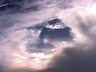 [within a circular opening in the clouds is a small segment of a rainbow. Although there is no arc, all the colors are present. The clouds are bright white]